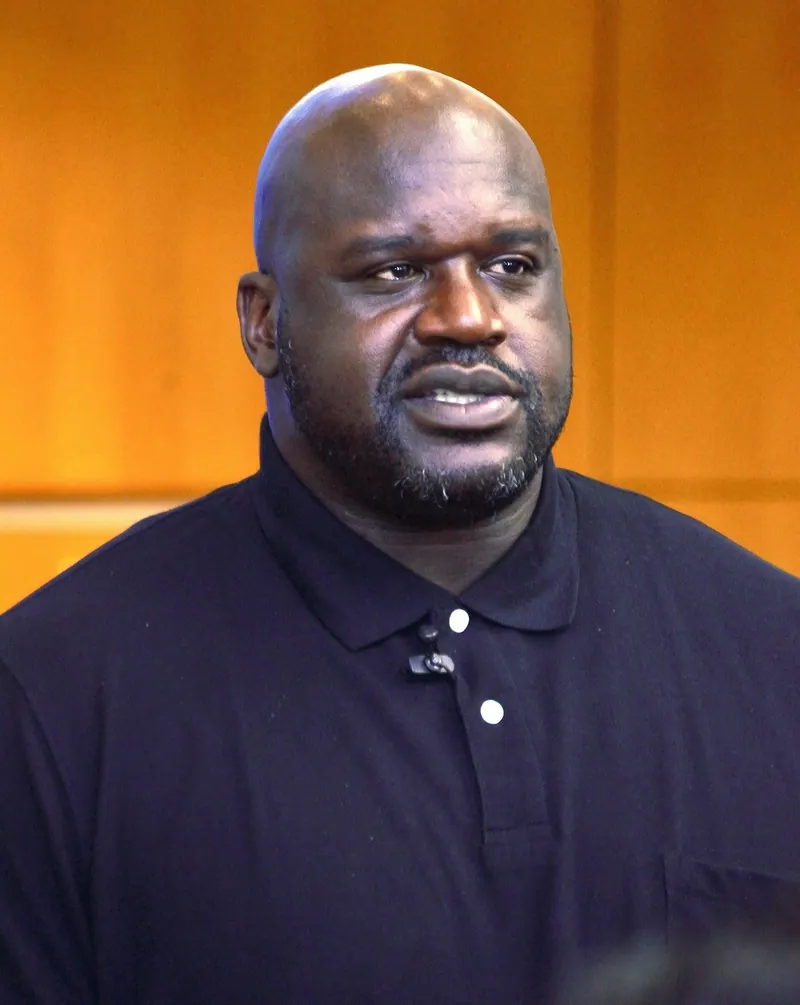 Close head-and-shoulders portrait of Shaquille O'Neal in a black polo shirt, speaking at a Dell community engagement (CE Day) event, warm wood-paneled background.