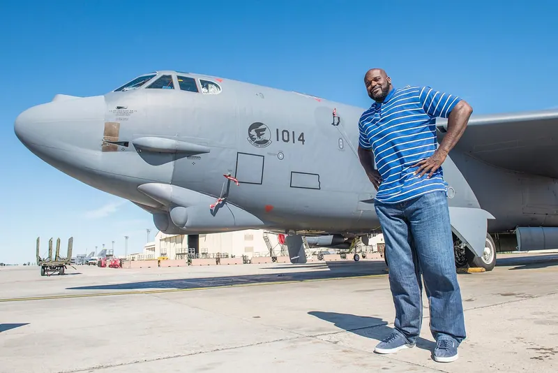 Shaquille O'Neal poses full-length beside the nose of a B-52H Stratofortress bomber under a blue sky at Minot Air Force Base, North Dakota, during a visit to speak with Airmen.