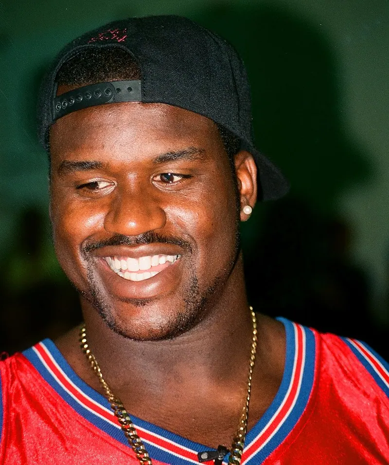 Close-up portrait of a young, broadly smiling Shaquille O'Neal wearing a backwards cap and chain necklace, taken in Washington, D.C. during his Lakers years.